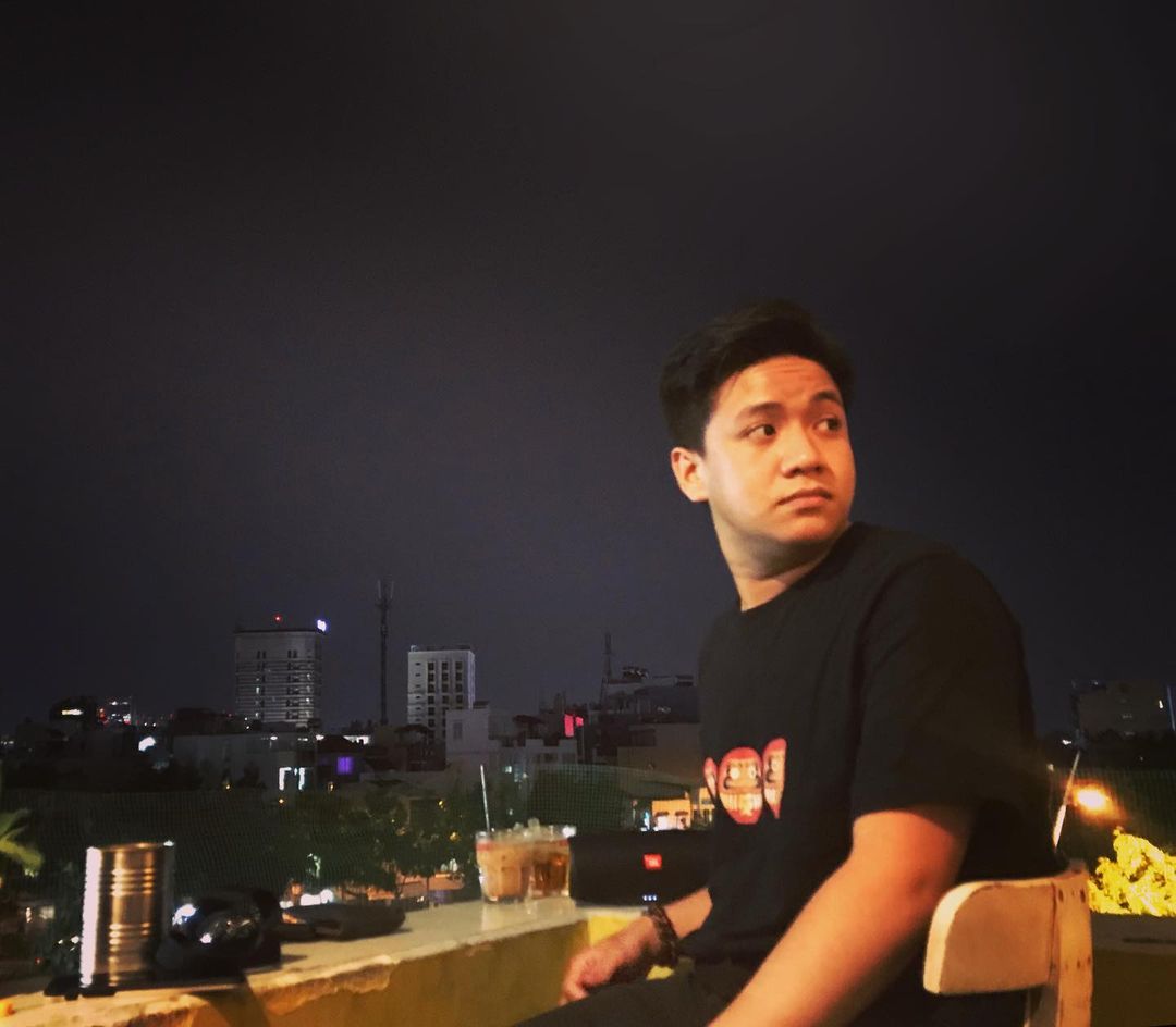 A rooftop at night, a figure looking off-frame with the lit city behind and a cold drink on the ledge.